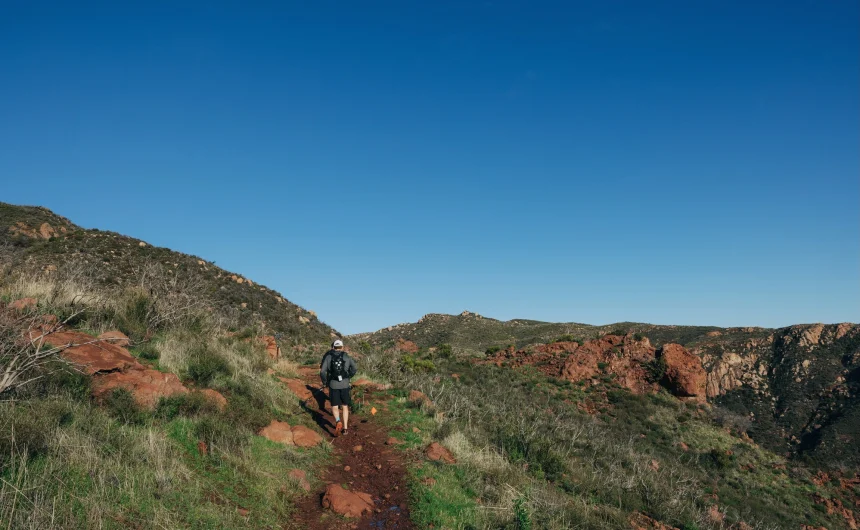 Hiker with backpack walking along a red dirt trail through a rocky desert landscape under a clear blue sky.