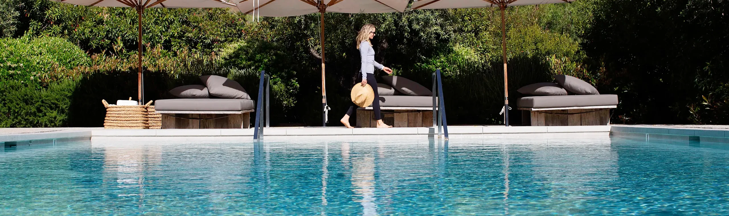 Woman walking barefoot along a poolside row of lounge chairs under large white umbrellas, holding a wide-brimmed hat amid lush greenery.