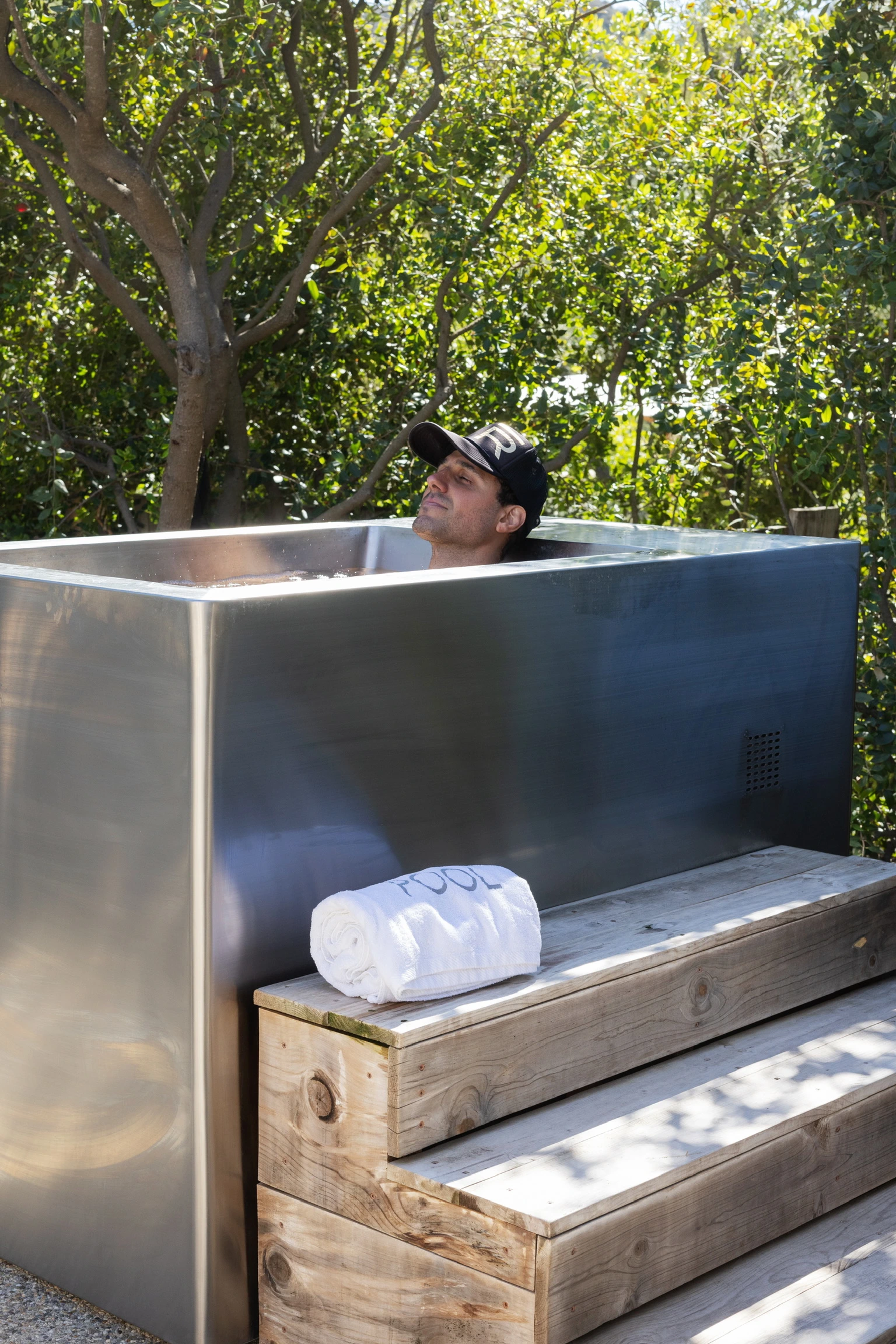 Man relaxing in a metal outdoor tub amid trees, with a towel on the wooden steps nearby.