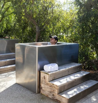 Man in a cap relaxing inside a stainless outdoor shower enclosure surrounded by wooden steps and trees on a sunny day.