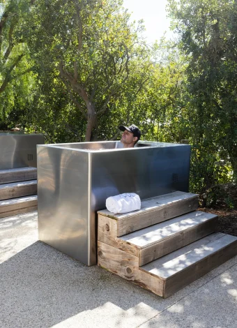 Man in a cap relaxing inside a stainless outdoor shower enclosure surrounded by wooden steps and trees on a sunny day.