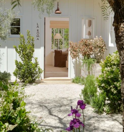 White cottage with an open doorway labeled LA ROMA; interior lamp and seating visible, gravel path and surrounding green plants outside.