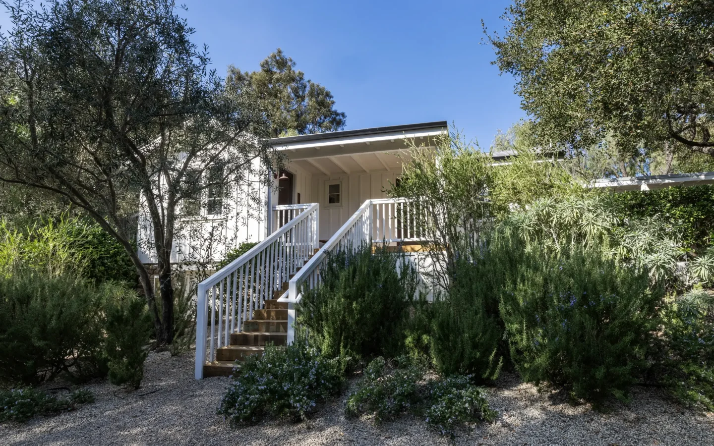White house entrance framed by lush shrubs and trees, wooden stairs and railing leading to a covered porch under a clear blue sky.