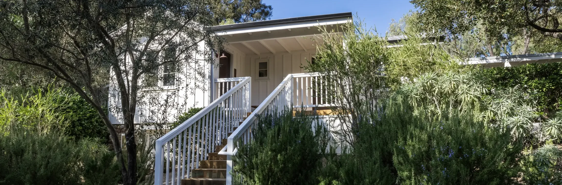 White house entrance framed by lush shrubs and trees, wooden stairs and railing leading to a covered porch under a clear blue sky.