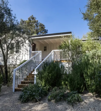 White house entrance framed by lush shrubs and trees, wooden stairs and railing leading to a covered porch under a clear blue sky.