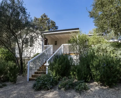 White house entrance framed by lush shrubs and trees, wooden stairs and railing leading to a covered porch under a clear blue sky.