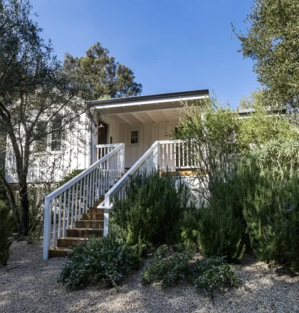 White house entrance framed by lush shrubs and trees, wooden stairs and railing leading to a covered porch under a clear blue sky.