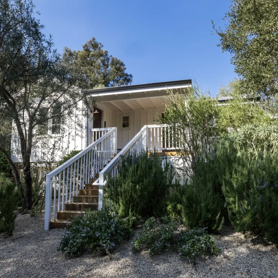 White house entrance framed by lush shrubs and trees, wooden stairs and railing leading to a covered porch under a clear blue sky.