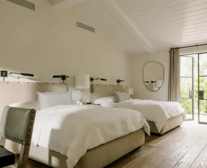 Bright hotel bedroom with two twin beds and beige upholstered headboards, lamps, and a large glass door to outdoors.