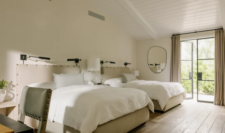 Bright hotel bedroom with two twin beds and beige upholstered headboards, lamps, and a large glass door to outdoors.