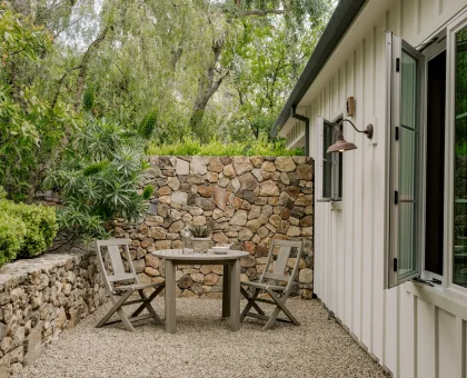 Outdoor courtyard with a round wooden table and four chairs, gravel ground, stone wall, and lush greenery beside a white house with open glass doors nearby.