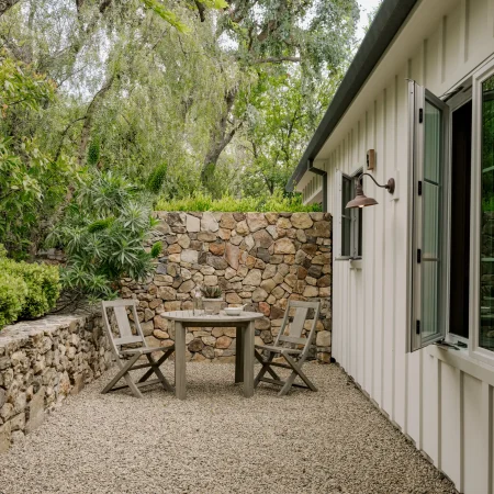 Outdoor courtyard with a round wooden table and four chairs, gravel ground, stone wall, and lush greenery beside a white house with open glass doors nearby.