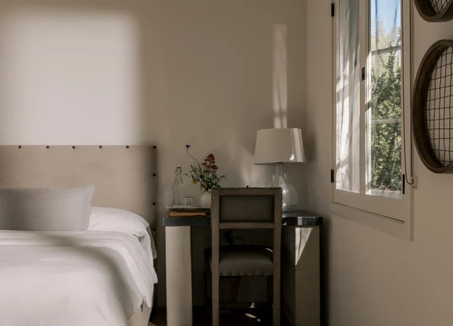 Bright bedroom corner with white bedding, a small wooden desk, lamp, and a window with a woven shade letting in sunlight.