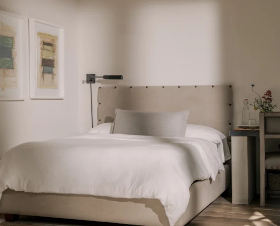 Cozy neutral bedroom with a beige upholstered bed, white linens, and a side table with flowers on the right.