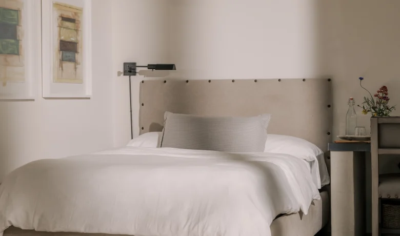 Cozy neutral bedroom with a beige upholstered bed, white linens, and a side table with flowers on the right.