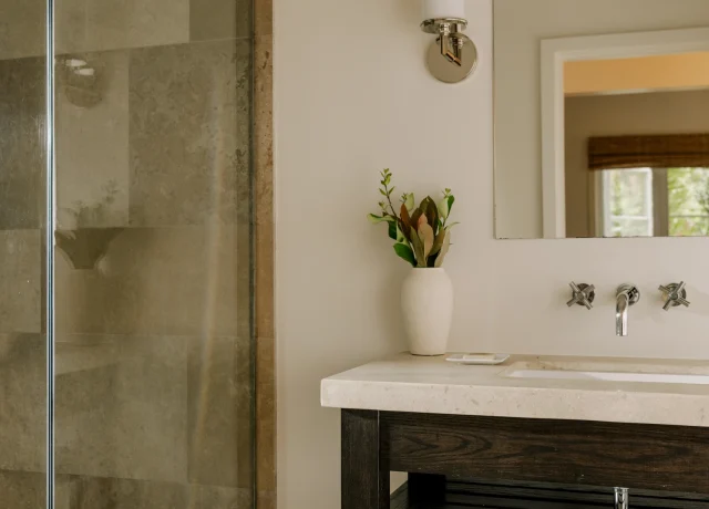 Bathroom area with glass-enclosed shower on the left, a wall light and mirror on the right, and a dark wood vanity with a vase of greenery on the counter.