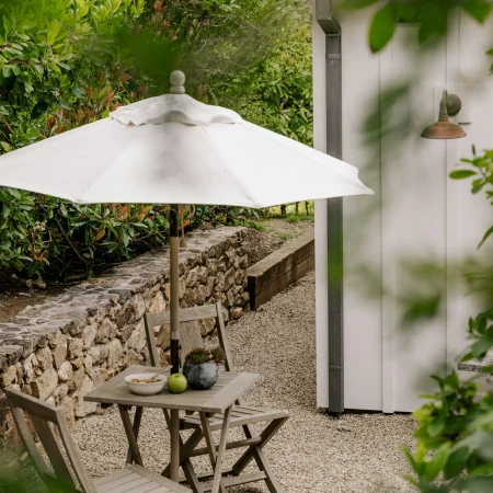 Outdoor gravel dining area with a round white umbrella, wooden table and chairs beside a stone wall and greenery