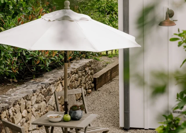 Outdoor gravel dining area with a round white umbrella, wooden table and chairs beside a stone wall and greenery
