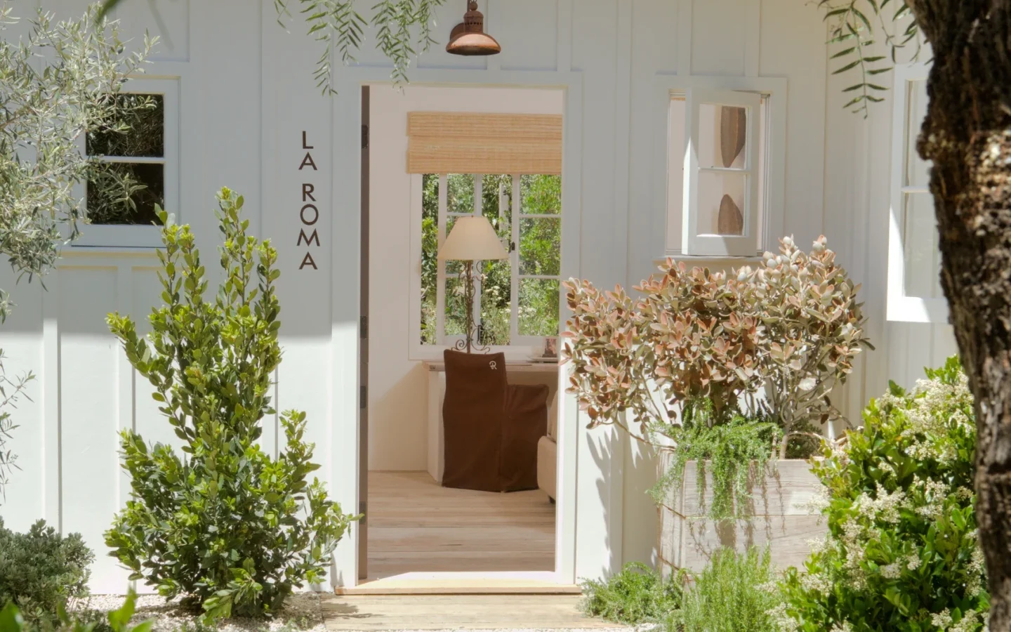 White doorway framed by lush greenery and hanging branches, with 'LA ROMA' signage on the side wall and a brown chair visible inside.