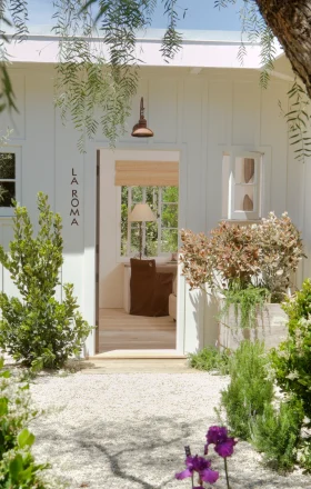 White doorway framed by lush greenery and hanging branches, with 'LA ROMA' signage on the side wall and a brown chair visible inside.