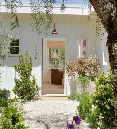 White doorway framed by lush greenery and hanging branches, with 'LA ROMA' signage on the side wall and a brown chair visible inside.