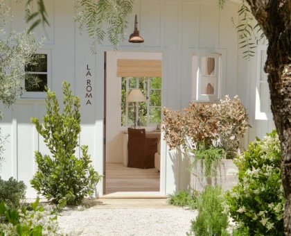 White doorway framed by lush greenery and hanging branches, with 'LA ROMA' signage on the side wall and a brown chair visible inside.