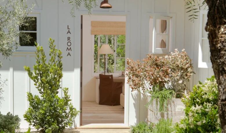 White doorway framed by lush greenery and hanging branches, with 'LA ROMA' signage on the side wall and a brown chair visible inside.