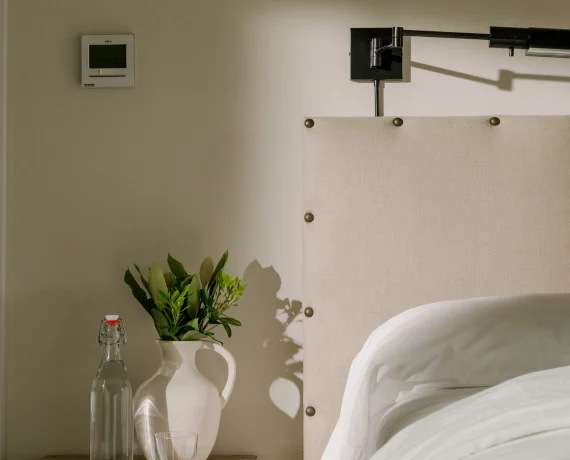 Bedroom bedside area with a wooden nightstand, glass bottle, ceramic pitcher, green plant, and a white bed with a beige upholstered headboard.