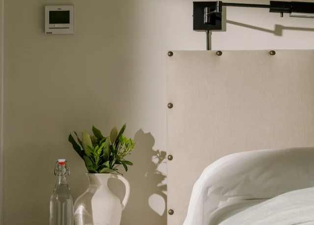 Bedroom bedside area with a wooden nightstand, glass bottle, ceramic pitcher, green plant, and a white bed with a beige upholstered headboard.