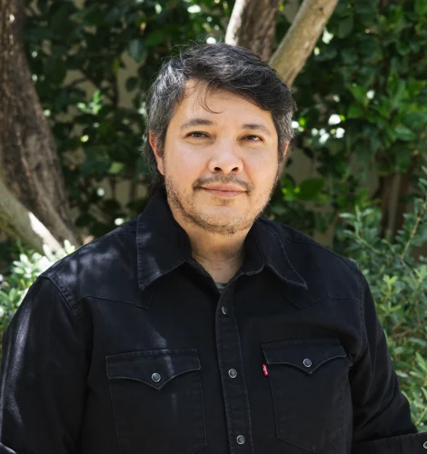 Man outdoors wearing a black button-down shirt, with green foliage and tree trunks in the background.