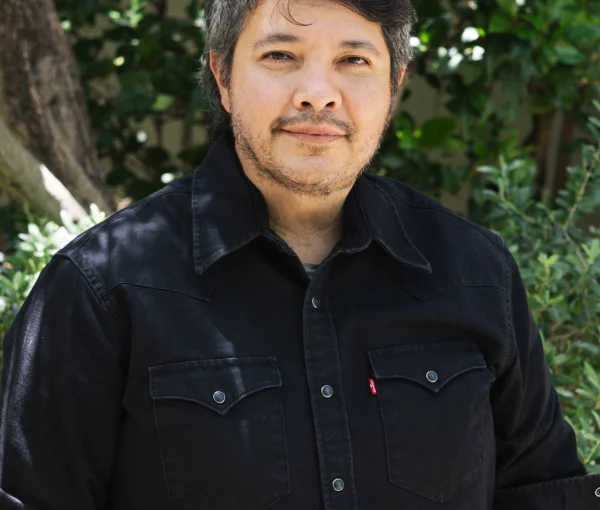 Man outdoors wearing a black button-down shirt, with green foliage and tree trunks in the background.