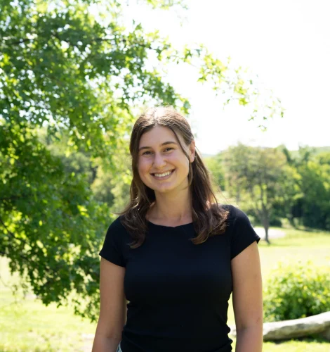 Smiling woman in a black T-shirt and denim shorts standing outdoors in a sunny park with trees in the background