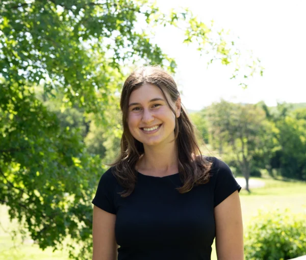 Smiling woman in a black T-shirt and denim shorts standing outdoors in a sunny park with trees in the background