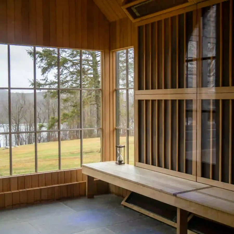 Sunlit wooden room with tall windows showing trees outside; a white table with a lantern sits along the wall.