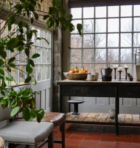 Sunlit indoor dining nook with large grid windows, potted plants, and a wooden table with a fruit bowl in the center of the room.