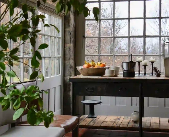 Sunlit indoor dining nook with large grid windows, potted plants, and a wooden table with a fruit bowl in the center of the room.