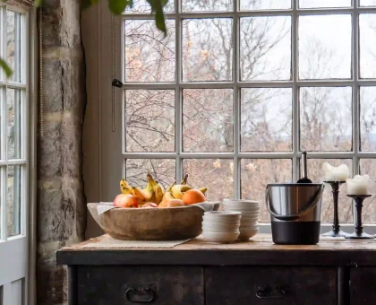 A wooden table with a fruit bowl, plates, and a stand mixer set by a large grid-window in a rustic kitchen.