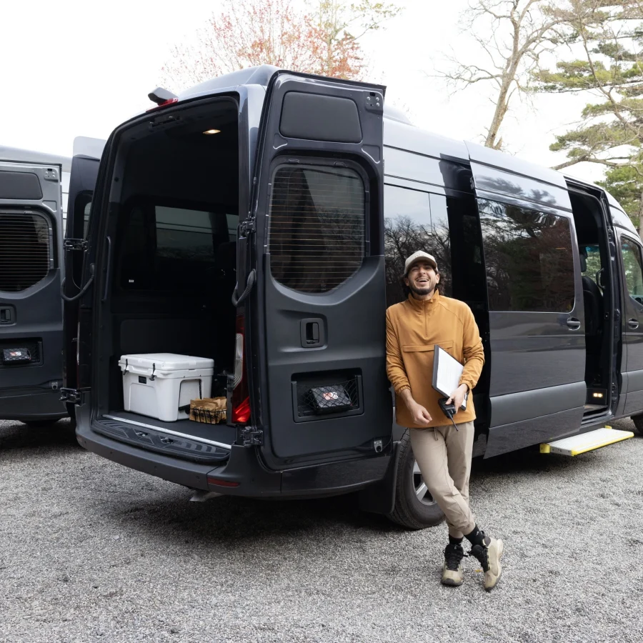 Man in a tan jacket exits a dark van with open rear doors, carrying gear in a parking lot.