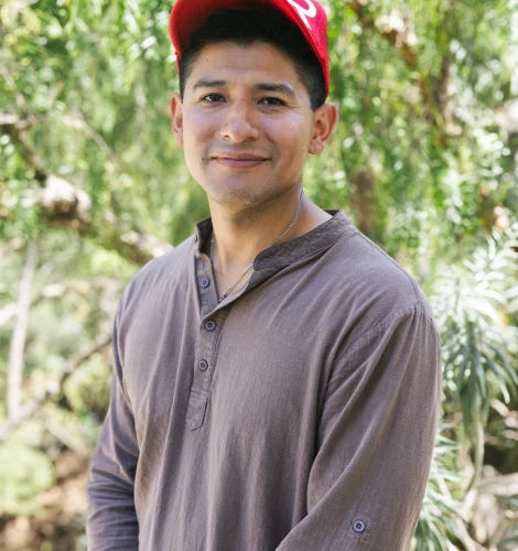 Smiling man wearing a red cap and brown shirt outdoors with green foliage behind him.