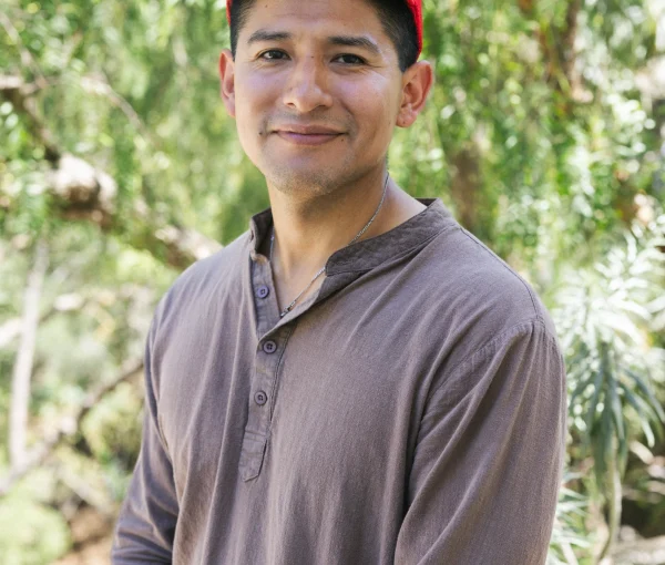 Smiling man wearing a red cap and brown shirt outdoors with green foliage behind him.