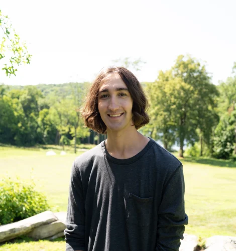 Young man with brown hair in a dark long-sleeve shirt smiling outdoors in a sunny park with trees behind him, camera angle waist up.