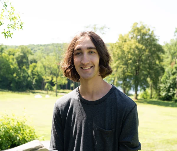Young man with brown hair in a dark long-sleeve shirt smiling outdoors in a sunny park with trees behind him, camera angle waist up.