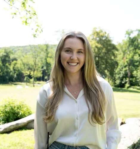 Smiling woman with long blonde hair standing outdoors in a sunny park, wearing a white blouse and jeans, with a radio clipped to her belt.