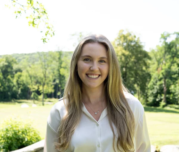 Smiling woman with long blonde hair standing outdoors in a sunny park, wearing a white blouse and jeans, with a radio clipped to her belt.