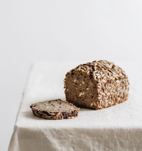 Seeded whole-grain loaf on a white table with a single slice showing dense crumb beside it on a light cloth.