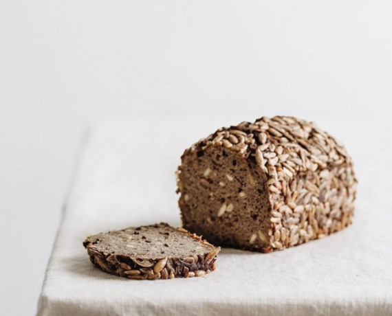 Seeded whole-grain loaf on a white table with a single slice showing dense crumb beside it on a light cloth.