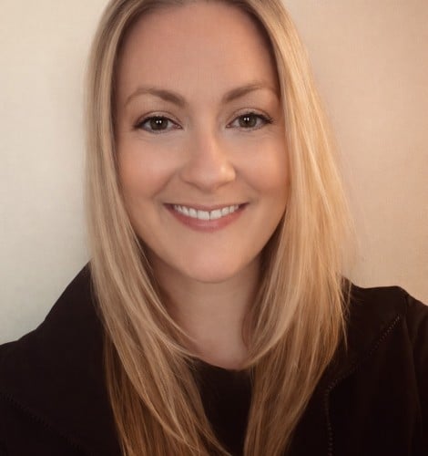 Smiling woman with shoulder-length blonde hair wearing a dark jacket against a light wall background.