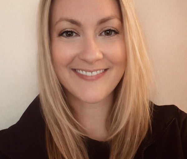 Smiling woman with shoulder-length blonde hair wearing a dark jacket against a light wall background.