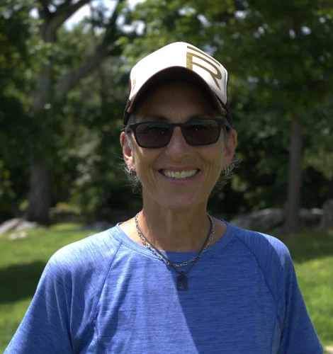 Smiling woman outdoors in a park wearing a beige cap, black sunglasses, a blue shirt, and a silver necklace.