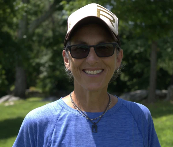 Smiling woman outdoors in a park wearing a beige cap, black sunglasses, a blue shirt, and a silver necklace.
